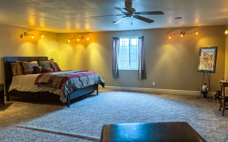 Spacious carpeted room featuring a ceiling fan, dual wall-mounted track lighting fixtures, and a large window with drapes