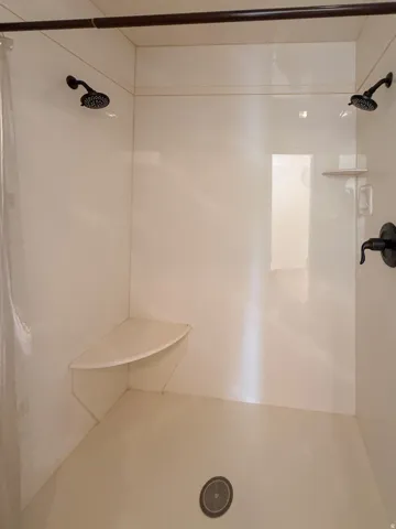 Shower enclosure featuring white paneling, dual shower heads with dark bronze finish, a built-in corner seat, and a built-in corner shelf