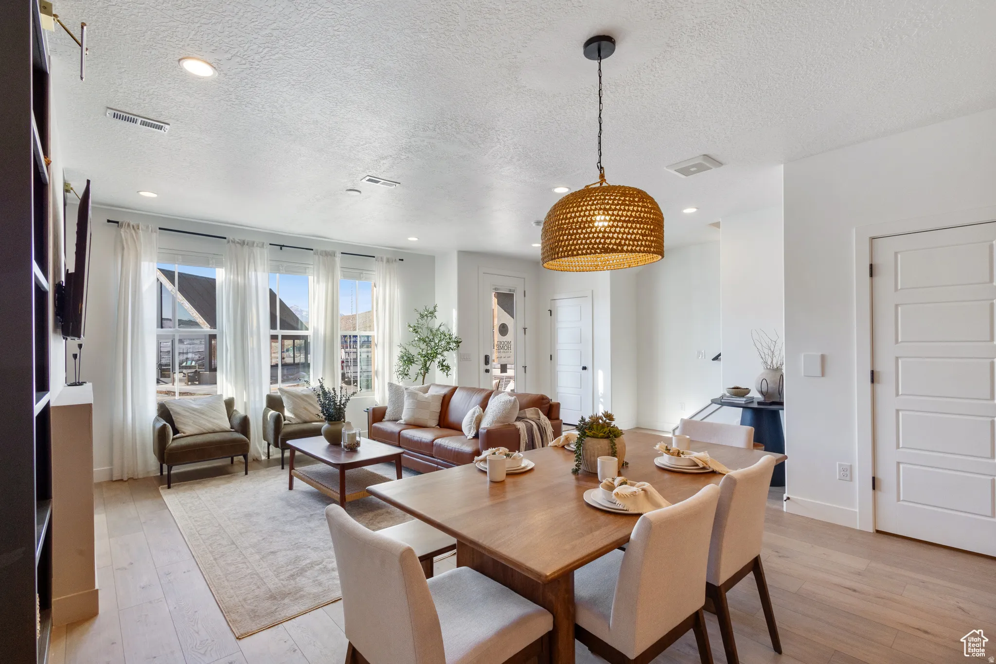 Open-concept living and dining area featuring light wood-finish flooring, recessed lighting, a woven pendant light fixture, and multiple large windows