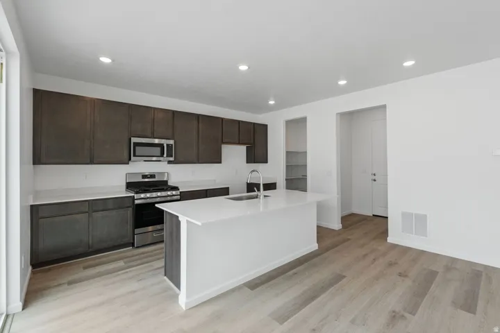 Kitchen featuring dark wood-finish cabinetry, stainless steel appliances, a center island with sink, and light wood-finish flooring
