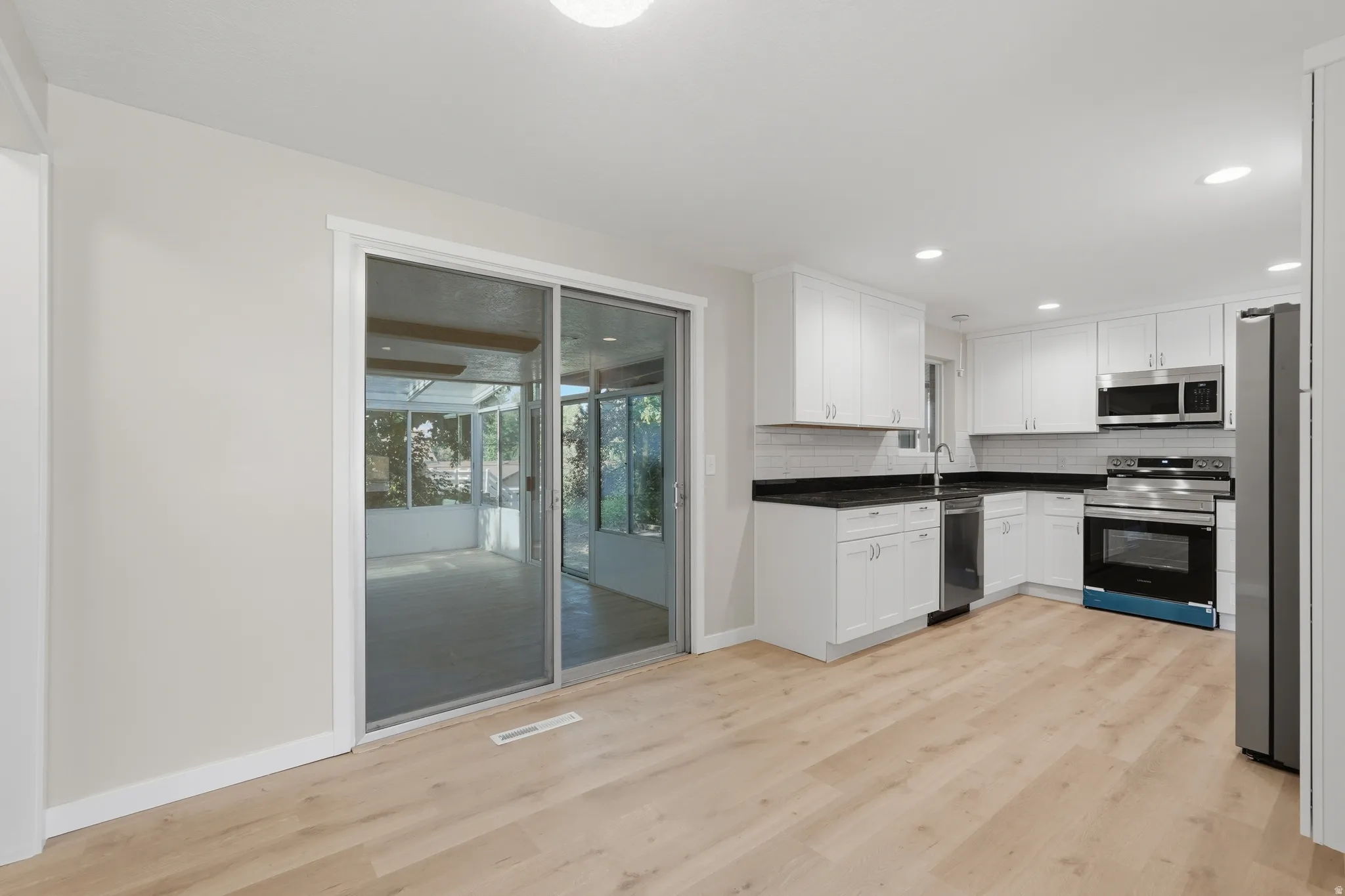 Open-concept kitchen featuring white shaker cabinetry, stainless steel appliances, black countertops, and a white subway tile backsplash