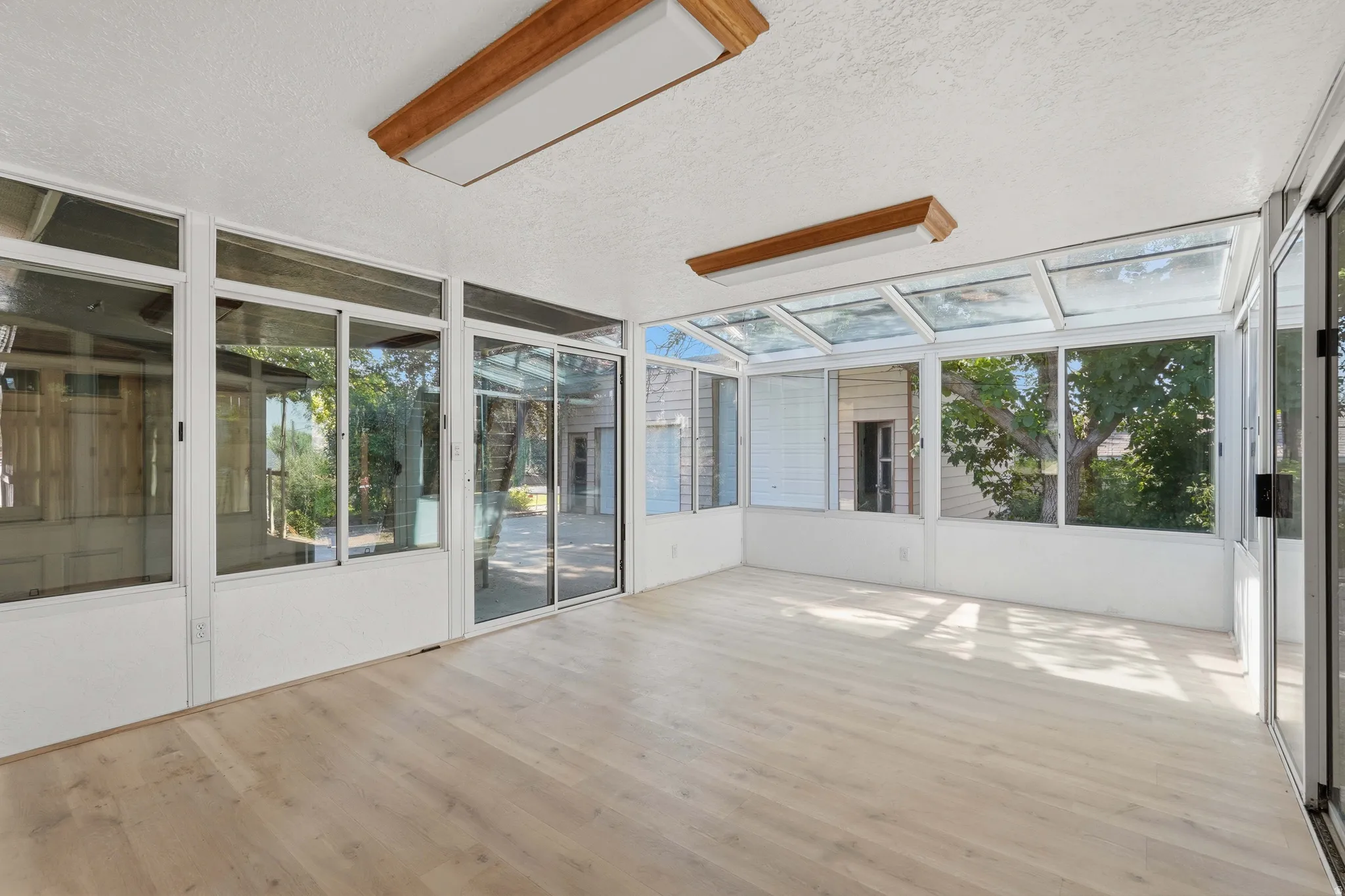 Sunroom featuring extensive glazing, a vaulted glass ceiling, wood-finish flooring, two rectangular fluorescent light fixtures, and sliding glass doors