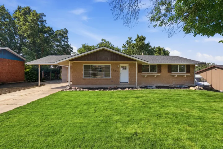 Brick-front residence featuring a covered carport, dark shingle roof, and a mature green lawn