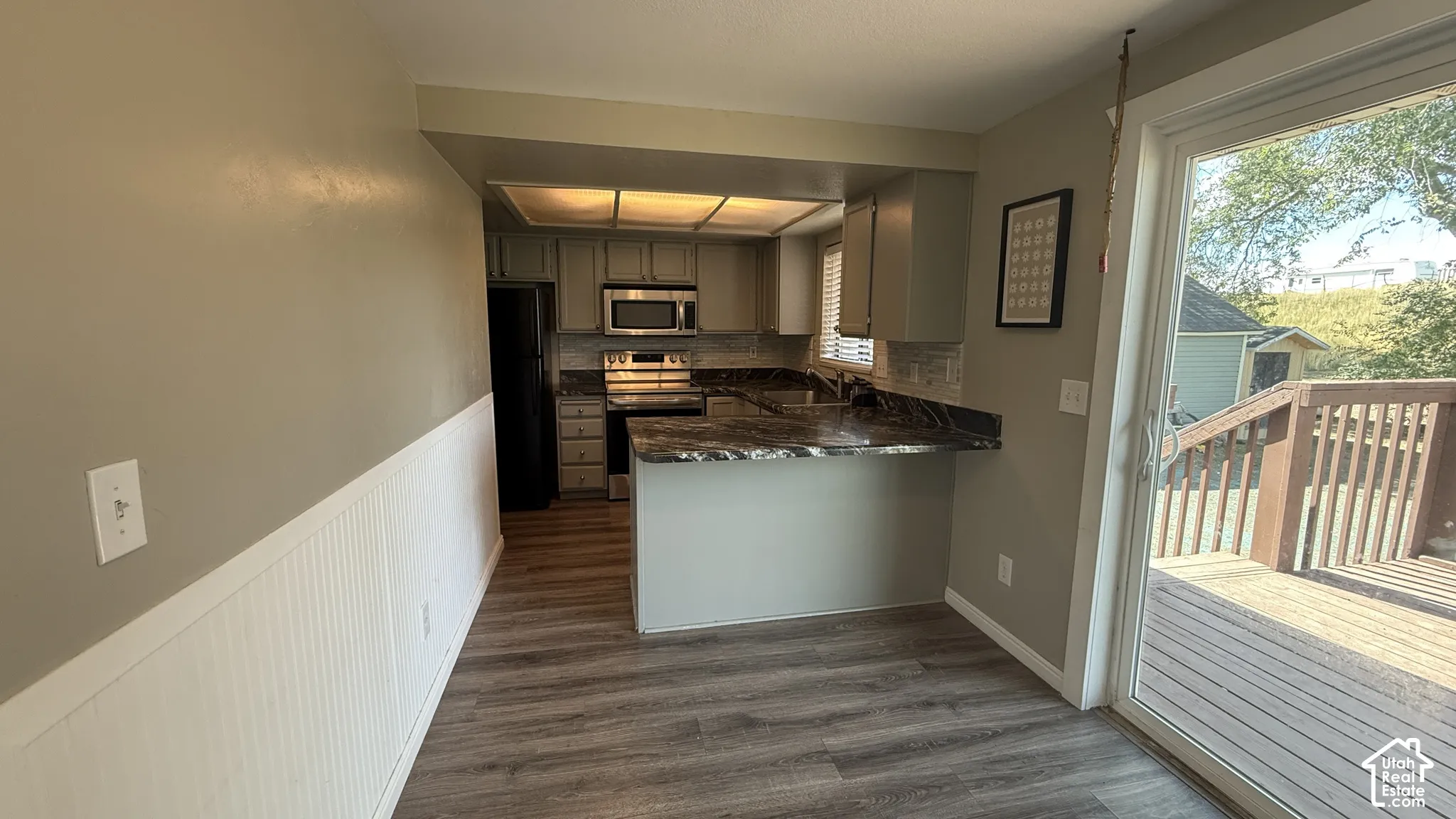 Open-concept kitchen and dining area featuring stainless steel appliances, dark wood-finish cabinetry with granite countertops, wood-finish flooring throughout, and abundant natural light from sliding glass doors opening to a covered deck