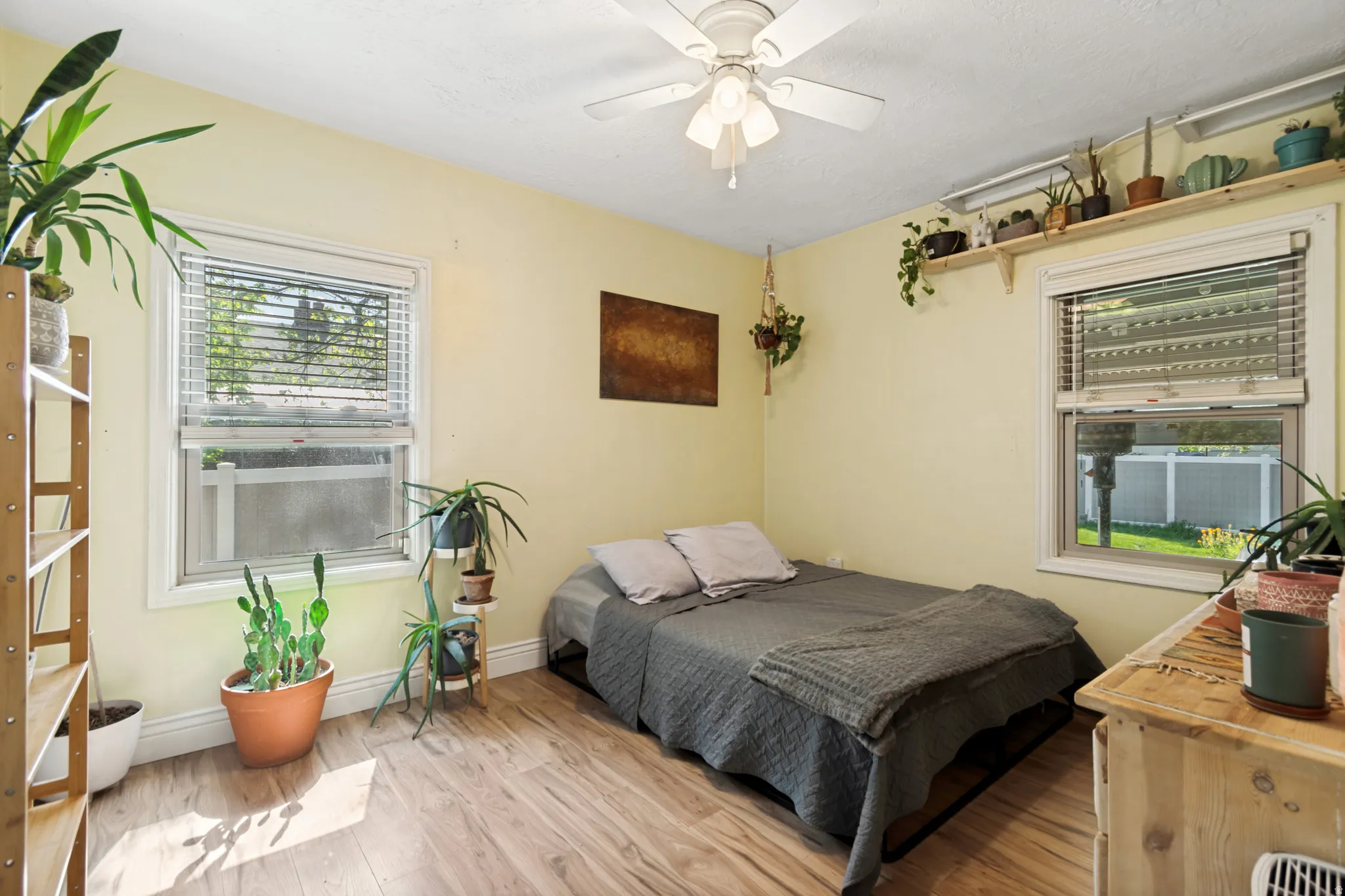 Bedroom featuring wood flooring, two window openings with blinds, a ceiling fan with integrated lighting, and a wall-mounted shelving unit