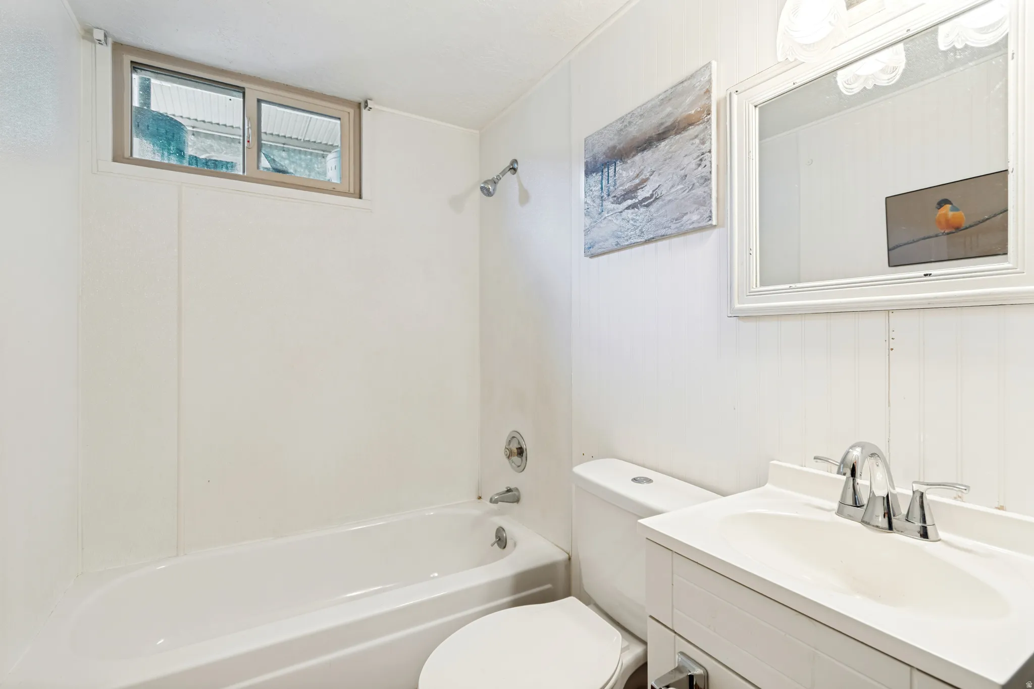 White bathroom featuring a bathtub with an integrated shower, a window with light-colored trim, a white vanity with an integrated sink, a chrome two-handle faucet, and vertical paneling on the wall