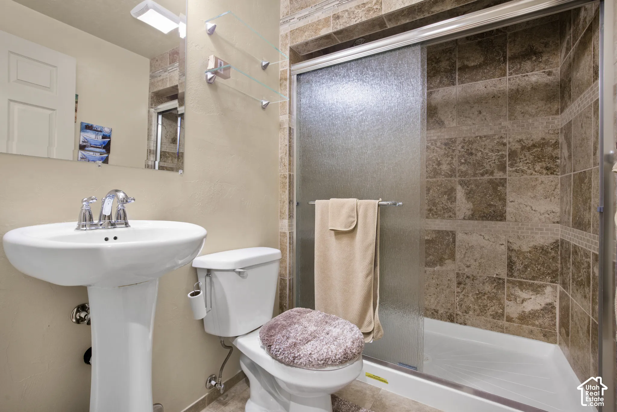 Bathroom featuring a pedestal sink, chrome faucet, wall-mounted mirror with integrated lighting, and a walk-in shower with stone tile surround and frosted glass sliding door