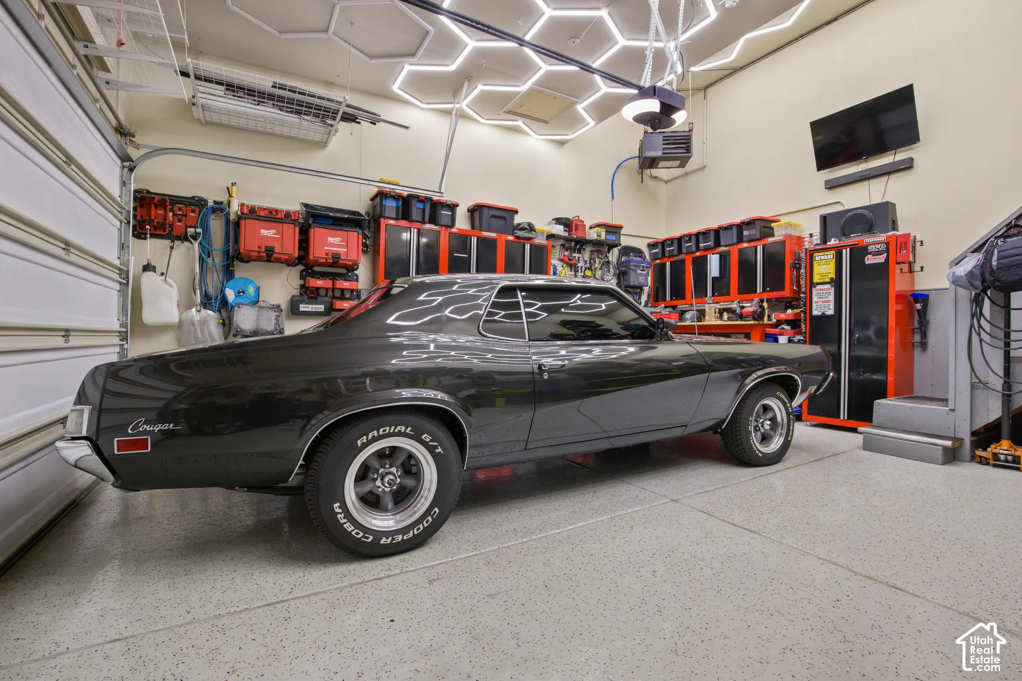 Spacious garage featuring a durable epoxy floor, integrated overhead storage, wall-mounted cabinetry, a built-in television, and bright overhead hexagonal lighting