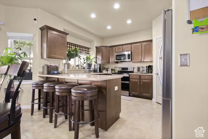 Kitchen featuring a breakfast bar with seating, recessed lighting, wood-finish cabinetry, stainless steel appliances, and light-toned tile flooring