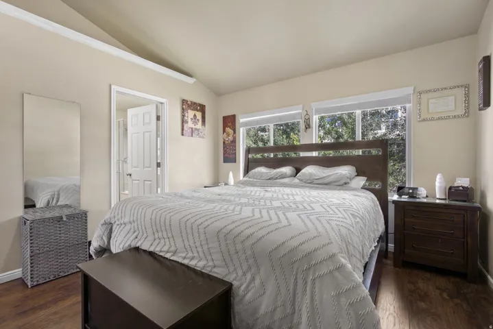 Vaulted ceiling bedroom with wood-finish flooring, two large windows, and a panel door leading to an adjacent room