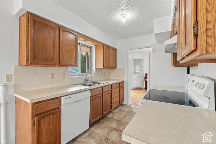 Galley kitchen featuring wood cabinetry, laminate countertops, white appliances including a dishwasher and range, tile flooring, and a window overlooking the exterior