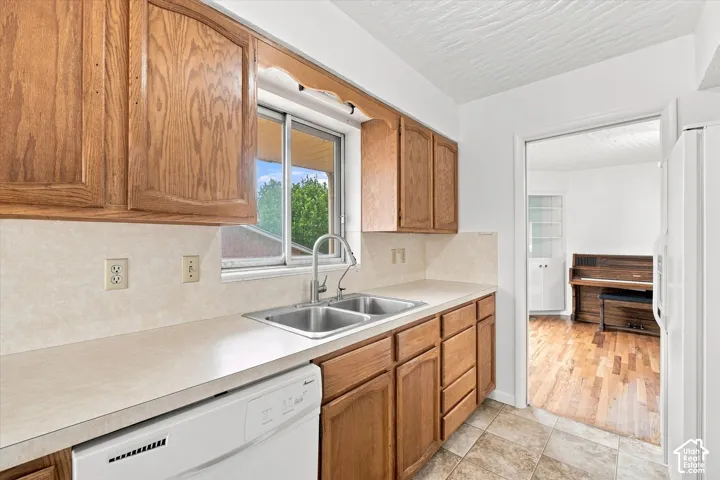 Kitchen featuring wood cabinetry, a double basin stainless steel sink with gooseneck faucet, light-toned countertop, and a built-in dishwasher