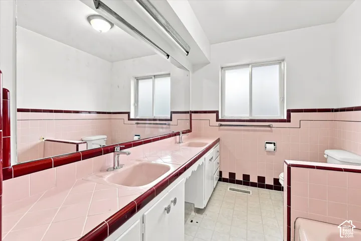 Bathroom featuring dual integrated sinks, an expansive vanity mirror, tiled walls with decorative trim, a window with privacy glass, and a built-in bathtub