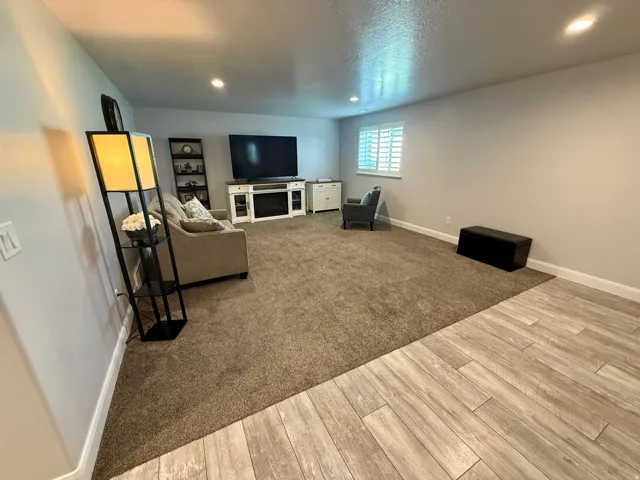Recessed lighting illuminates this versatile room, featuring a built-in media console with fireplace, a window with plantation shutters, and a combination of wood-finish flooring and plush carpeting