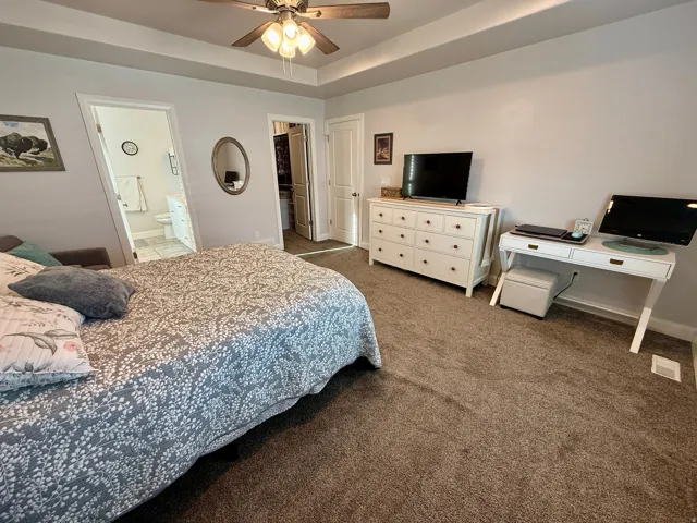 Carpeted room featuring a tray ceiling with a ceiling fan, light gray wall paint, and an en-suite bathroom with tiled flooring and a white vanity