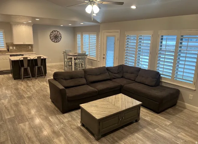 Open-concept living space featuring wood-finish flooring, a kitchen island with seating, recessed lighting, ceiling fan, and plantation shutters