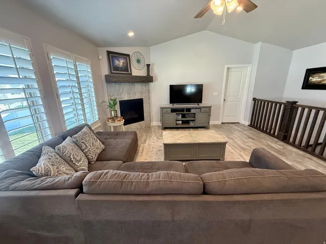 Living area featuring vaulted ceilings, a ceiling fan, a fireplace with a stone surround and wood mantle, wood-finish flooring, and white plantation shutters