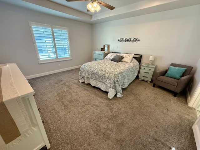Spacious bedroom featuring plush carpeting, a tray ceiling with a built-in ceiling fan, and a window with plantation shutters