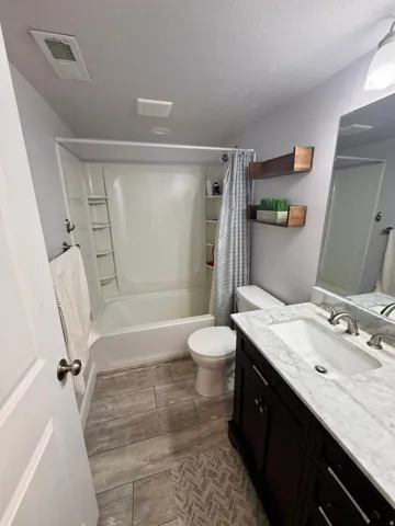 Bathroom featuring a white bathtub/shower combination, dark wood-finish vanity with a white countertop and integrated sink, brushed nickel fixtures, and wood-finish tile flooring
