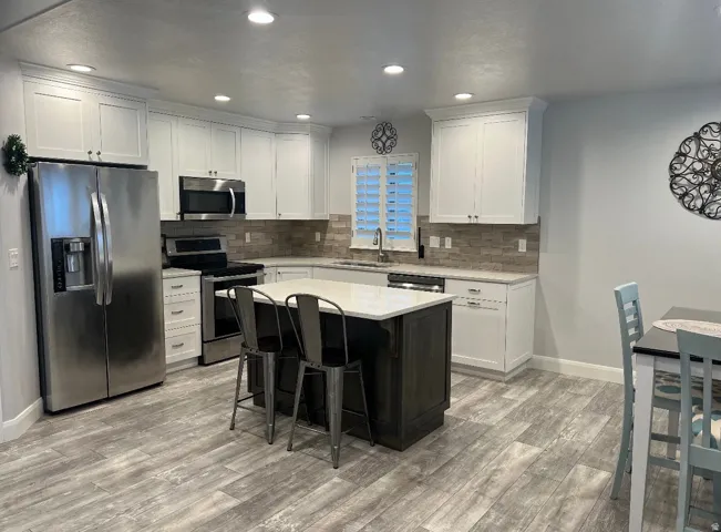 Modern kitchen featuring an island with seating, stainless steel appliances, white cabinetry, light-toned countertops, and wood-finish flooring
