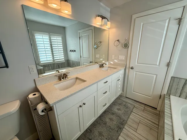 Bathroom featuring a double vanity with stone countertops and undermount sinks, an expansive wall-mounted mirror, white shaker-style cabinetry, and wood-look tile flooring