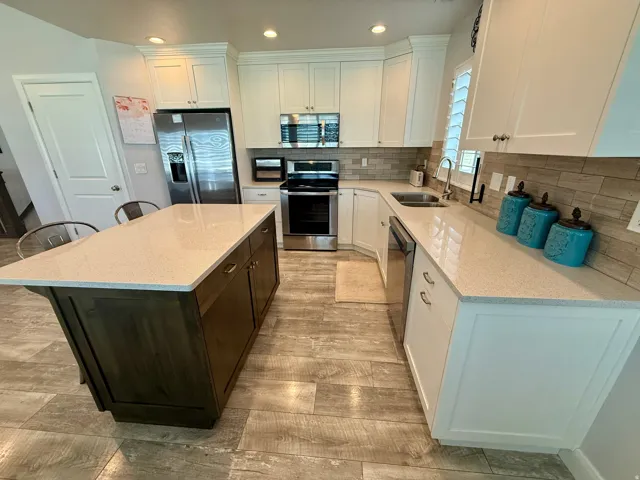 Spacious kitchen featuring an island with a light-toned countertop, wood-finish flooring, white cabinetry, stainless steel appliances, and a tiled backsplash