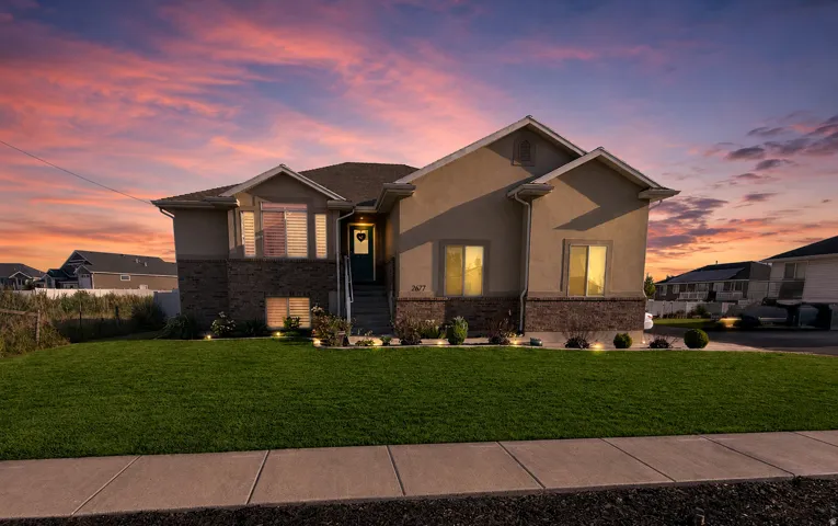 Contemporary residence featuring a stucco and stone facade, a gabled roofline, a manicured front lawn, and foundational landscaping with accent lighting