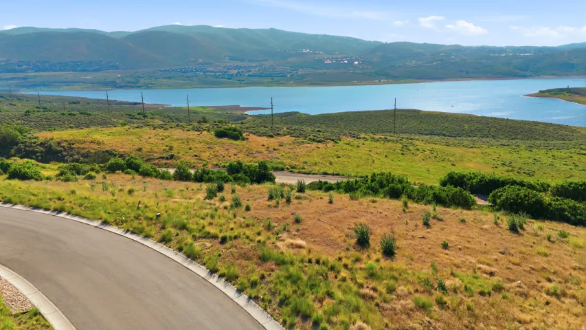 Paved road bordering an expanse of natural terrain, featuring varied vegetation, a prominent body of water, and distant mountain ranges under a clear sky