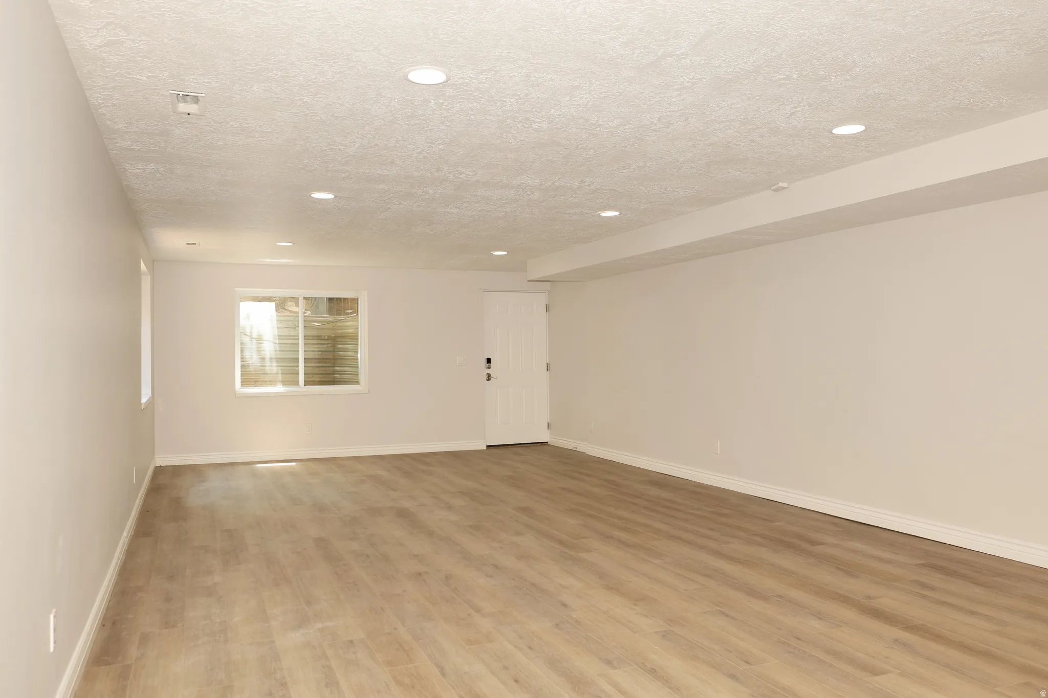 Spacious room with wood-finish flooring, recessed lighting, white walls, and a visible window well