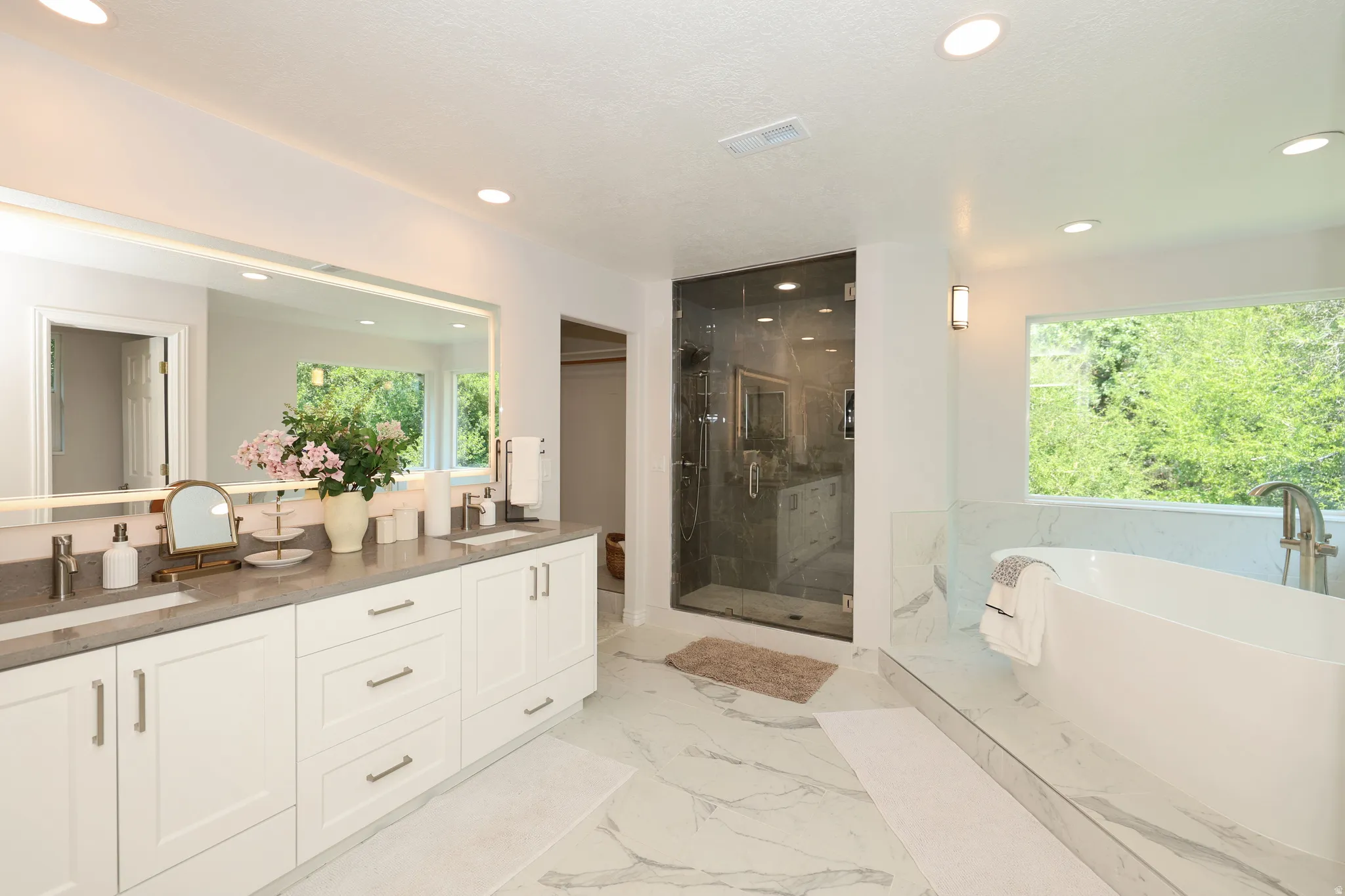 Spacious bathroom featuring a dual vanity with undermount sinks and stone countertops, a frameless glass shower enclosure, a freestanding soaking tub, and large format marble-look tile flooring