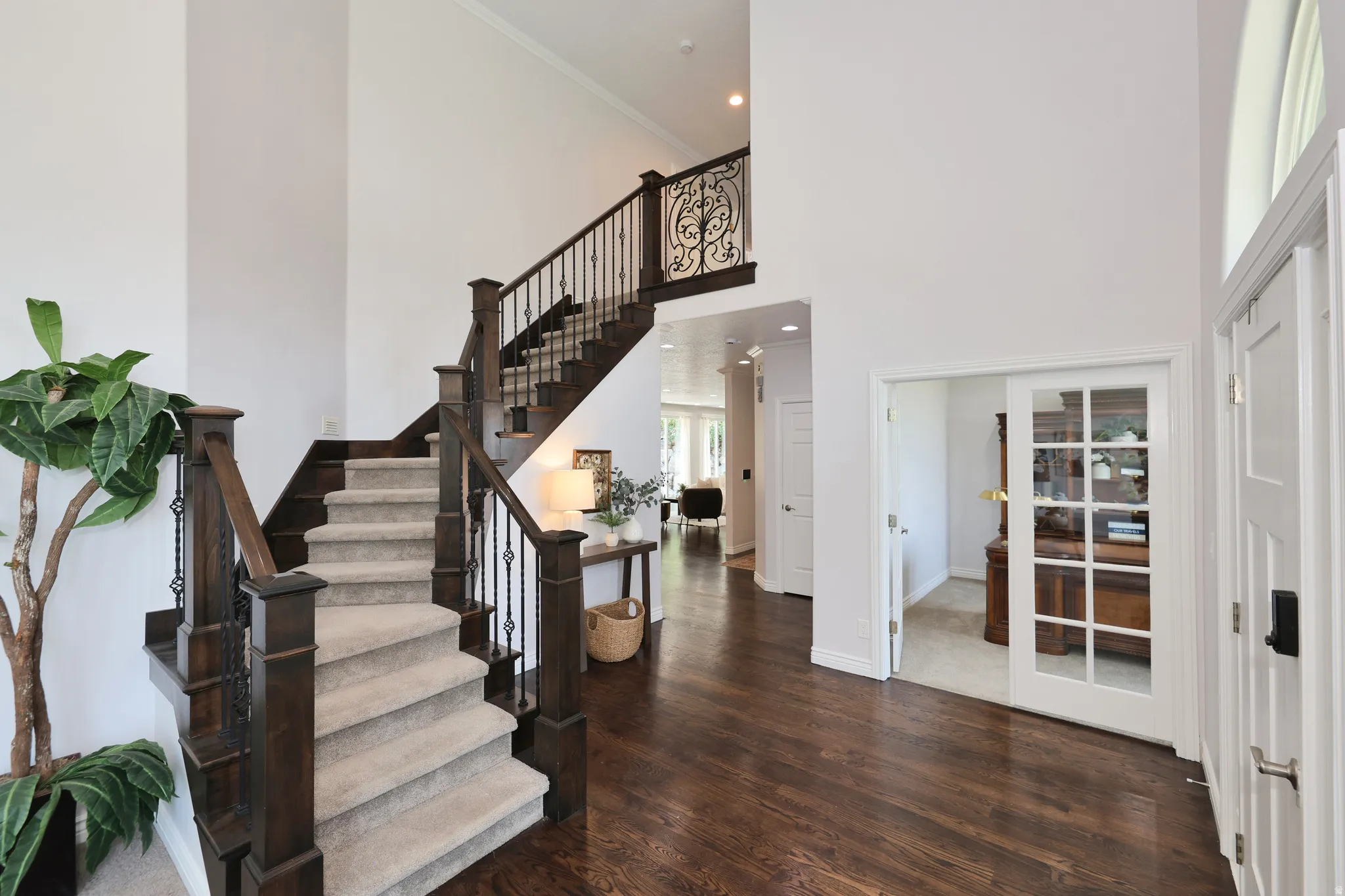 Two-story foyer featuring dark wood-finish flooring, a grand staircase with carpeted treads, dark wood newel posts, and wrought iron balusters, and a doorway with French doors