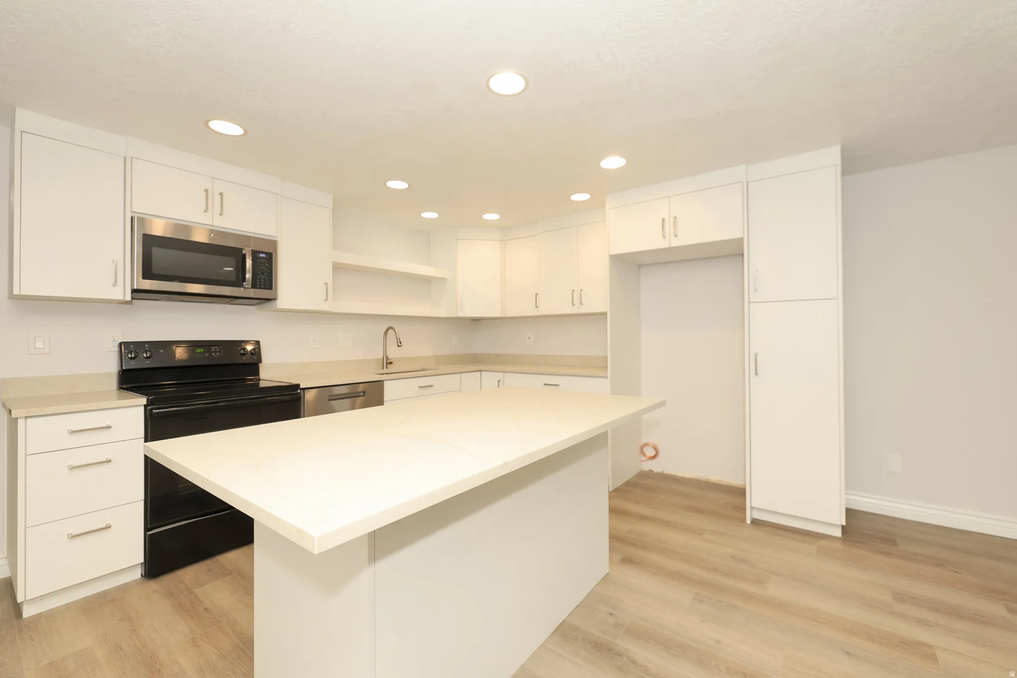 Modern kitchen featuring white cabinetry, light-toned countertops, stainless steel microwave, black electric range, and wood-finish flooring
