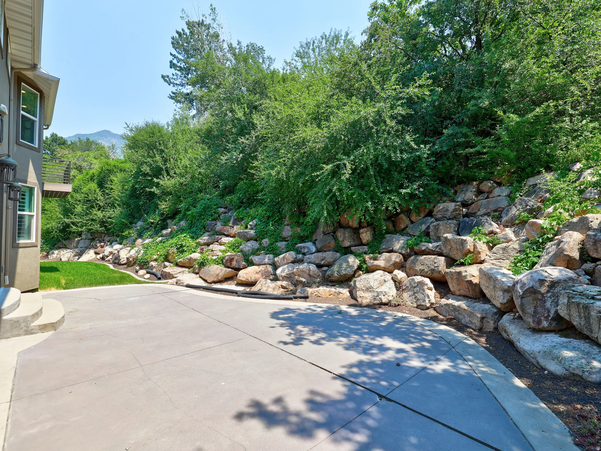 Spacious concrete patio with a tiered rock retaining wall and lush green landscaping