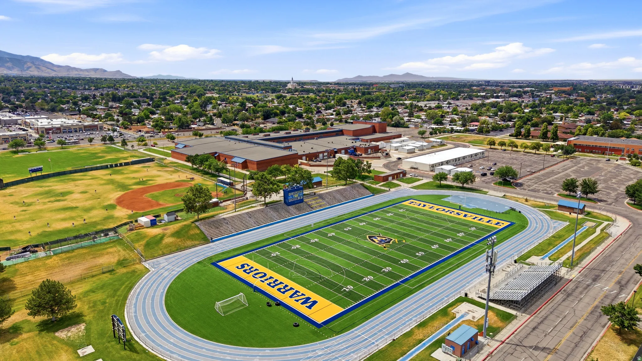 Aerial perspective showcasing a sports complex with a synthetic turf football field, a blue track, bleacher seating, a baseball diamond, and surrounding structures