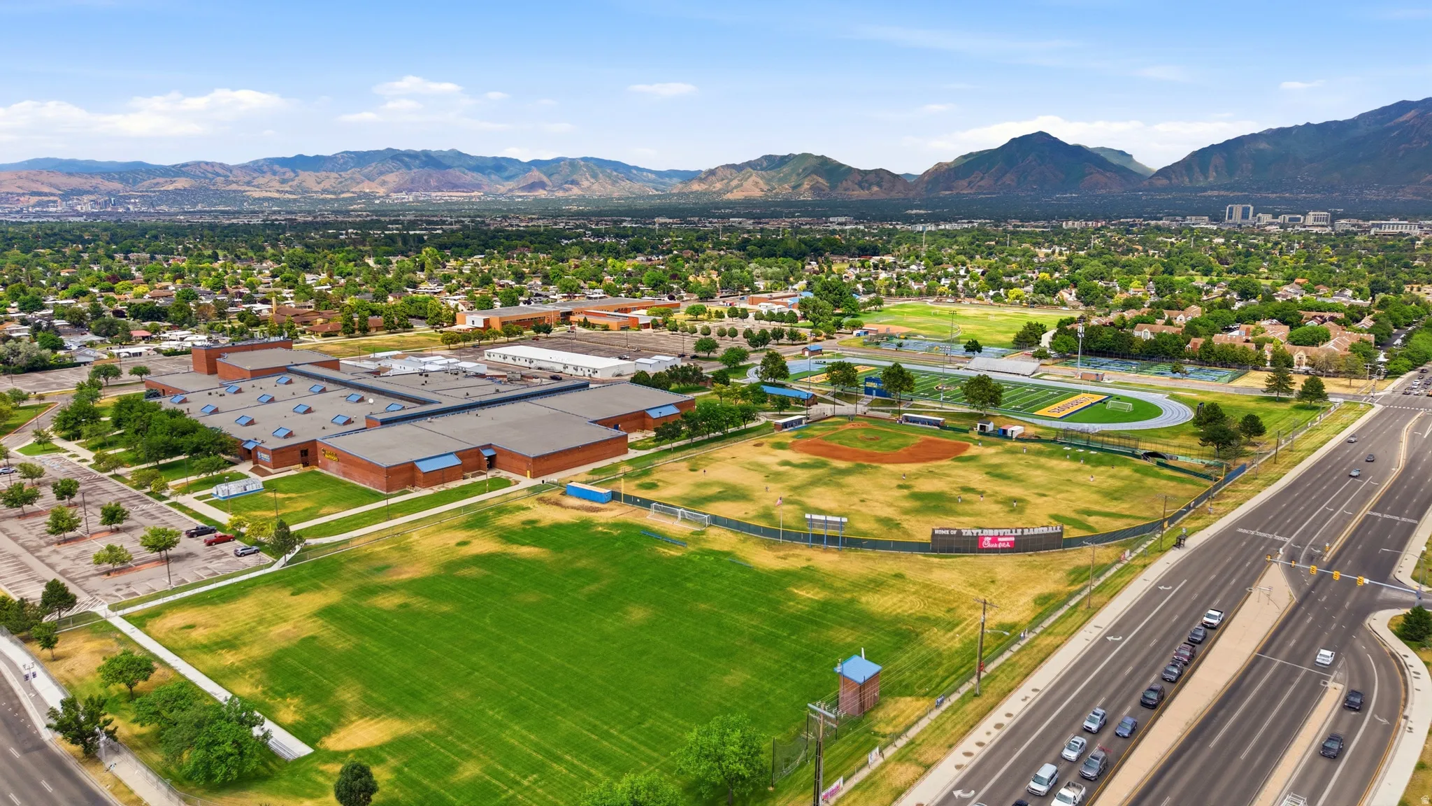 Expansive aerial capturing a large institutional building with multiple wings, featuring a substantial parking area and surrounded by green athletic fields including a baseball diamond and a running track