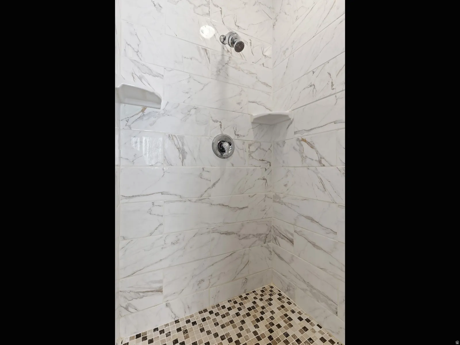 Shower stall featuring white marble-look subway tile walls with subtle gray veining, a mosaic tile floor in neutral tones, a built-in shower niche, and a chrome shower head and faucet