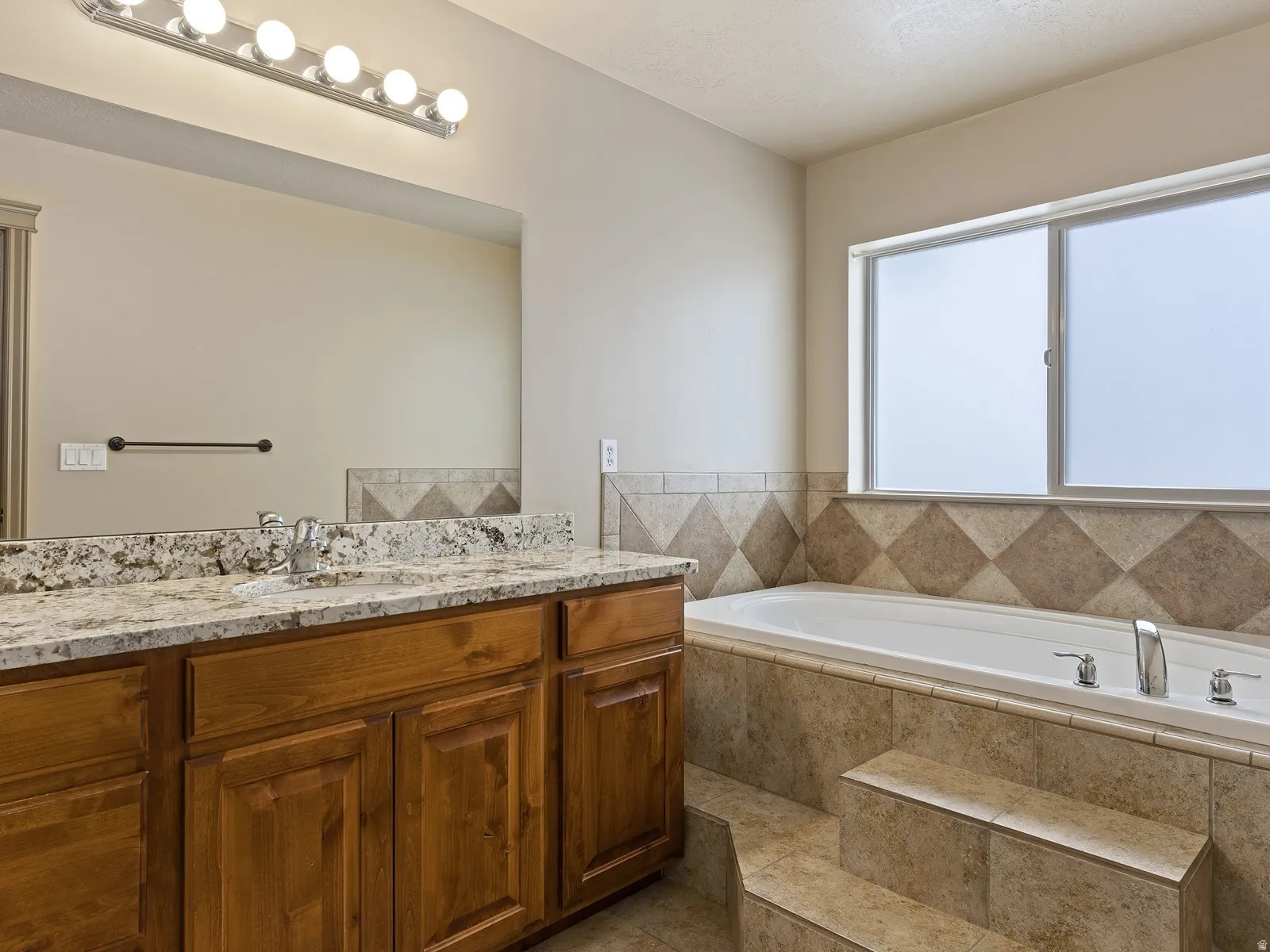Spacious bathroom featuring a built-in soaking tub with tiled surround, a large window, a dual-sink vanity with a stone-finish countertop, and a wide mirror with overhead lighting