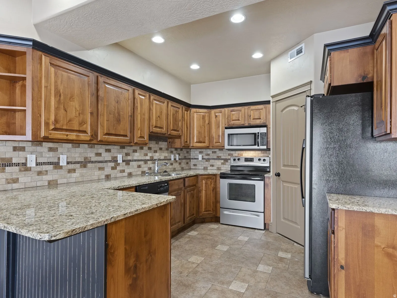 Kitchen featuring light wood-finish cabinetry with dark crown molding, a mosaic tile backsplash, granite-finish countertops, stainless steel appliances, and recessed lighting