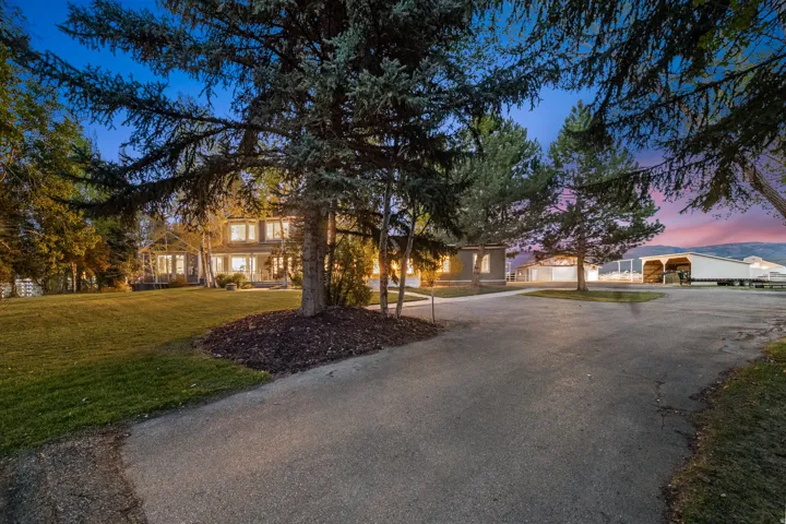Spacious property featuring a large residence with multiple windows, a broad paved driveway, extensive green lawn areas, mature evergreen trees, and distant mountains