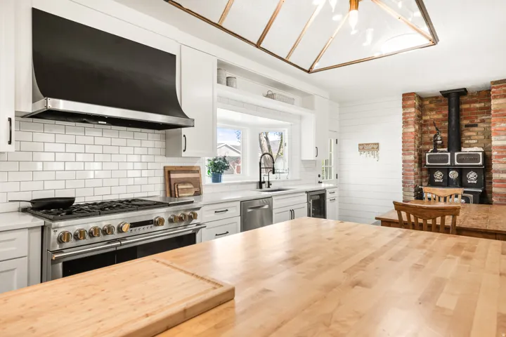 Spacious kitchen featuring a large wood butcher block island, a professional-grade stainless steel range with a custom black hood, white subway tile backsplash, white shaker cabinetry, and a brick accent wall with a wood-burning stove
