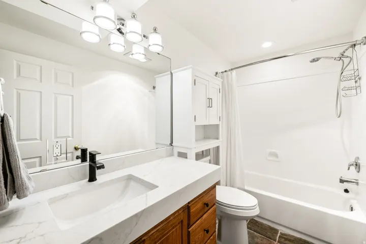 Bathroom featuring a white bathtub with a shower system, a wood-finish vanity with a white countertop and integrated sink, a matte black faucet, and a wall-mounted mirror