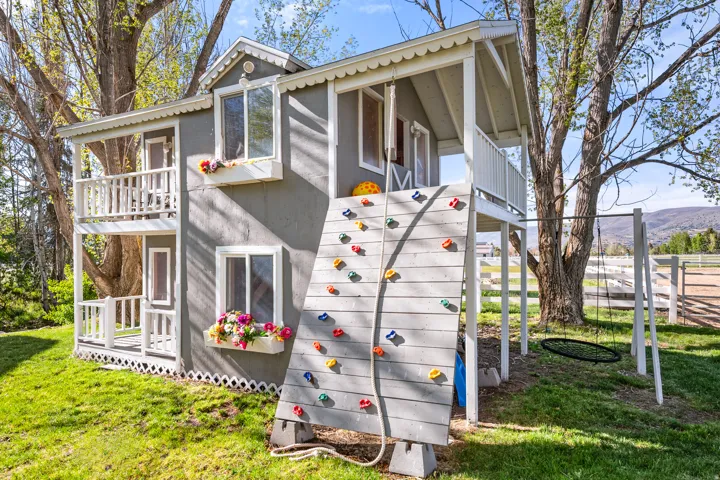 Custom two-story playhouse featuring multiple windows, a dual-level porch with railings, an integrated rock climbing wall, and a swing set