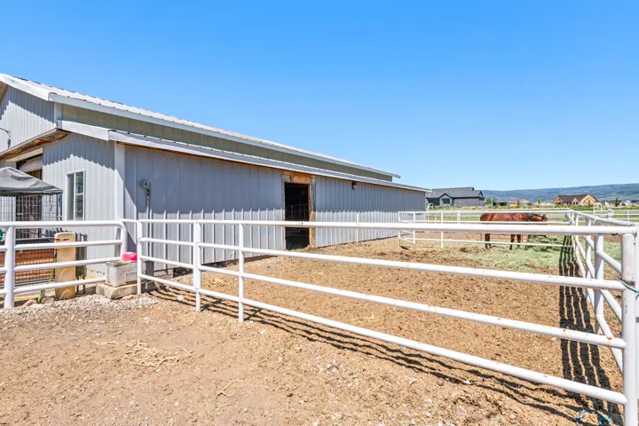 Gray metal-sided barn with a white metal roof, featuring multiple enclosed paddocks with white metal fencing