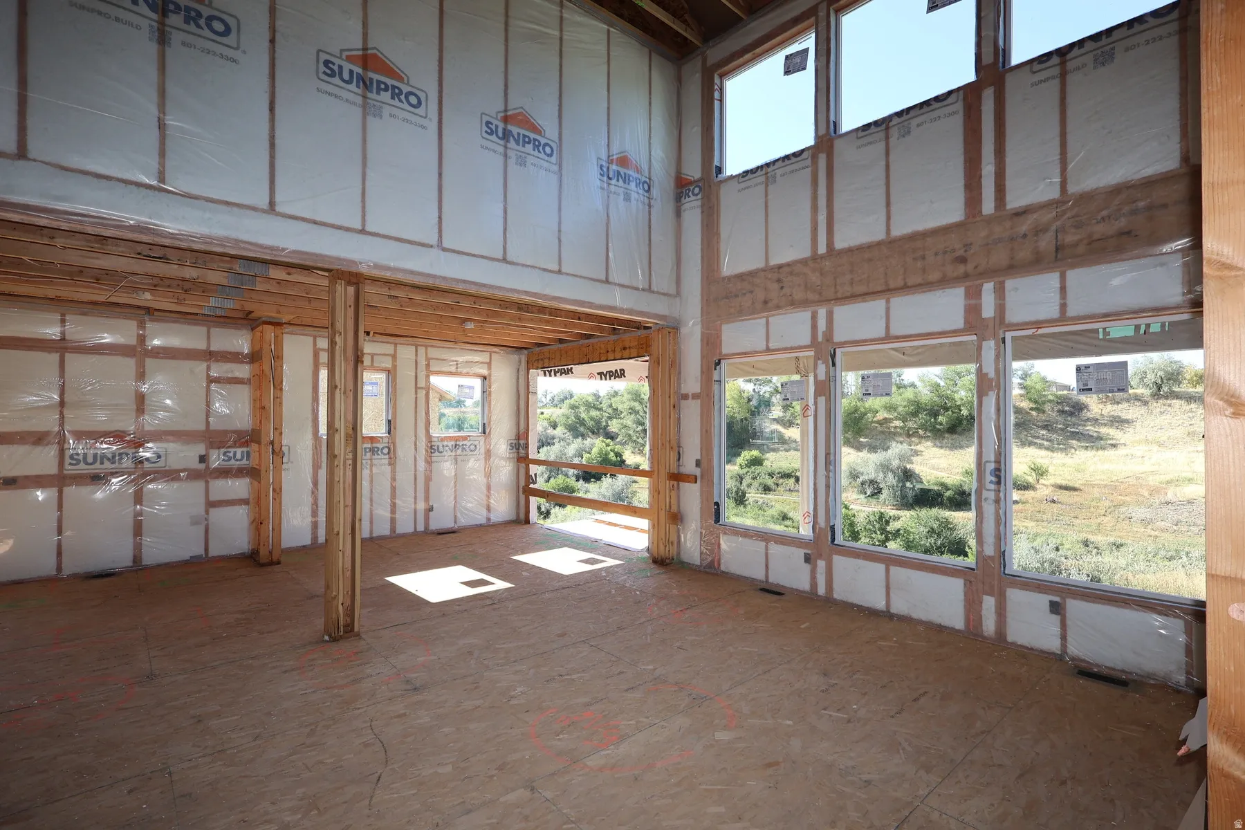 Expansive interior space featuring structural wood framing, a two-story window arrangement, and plywood subflooring