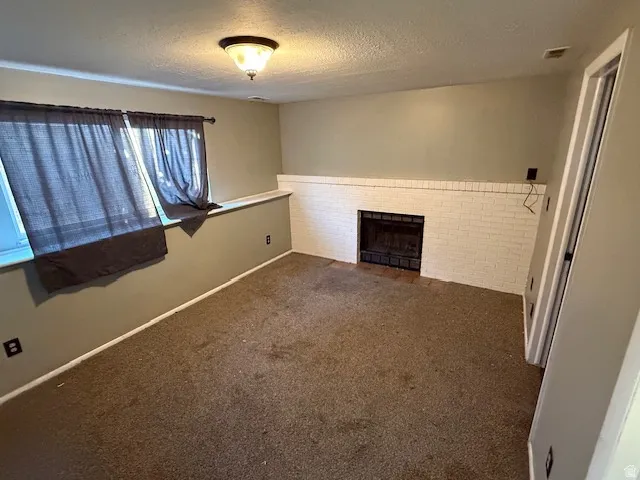 Room featuring a white painted brick fireplace with a black grate, two windows, carpet flooring, and a ceiling-mounted light fixture