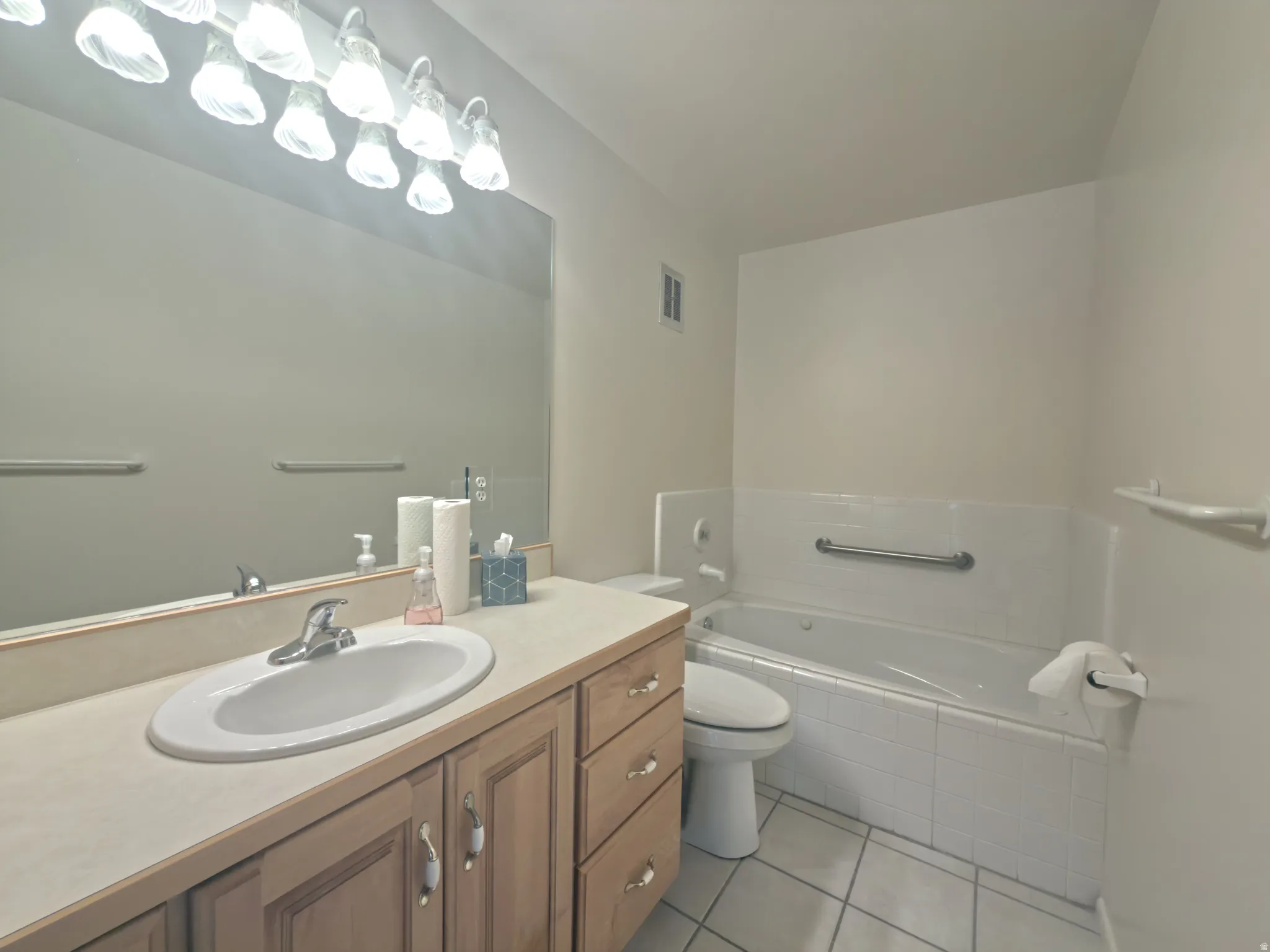 Bathroom featuring a light wood-finish vanity with an integrated sink and chrome faucet, a large wall-mounted mirror, overhead track lighting, a tiled tub surround, and a grab bar in the tub area