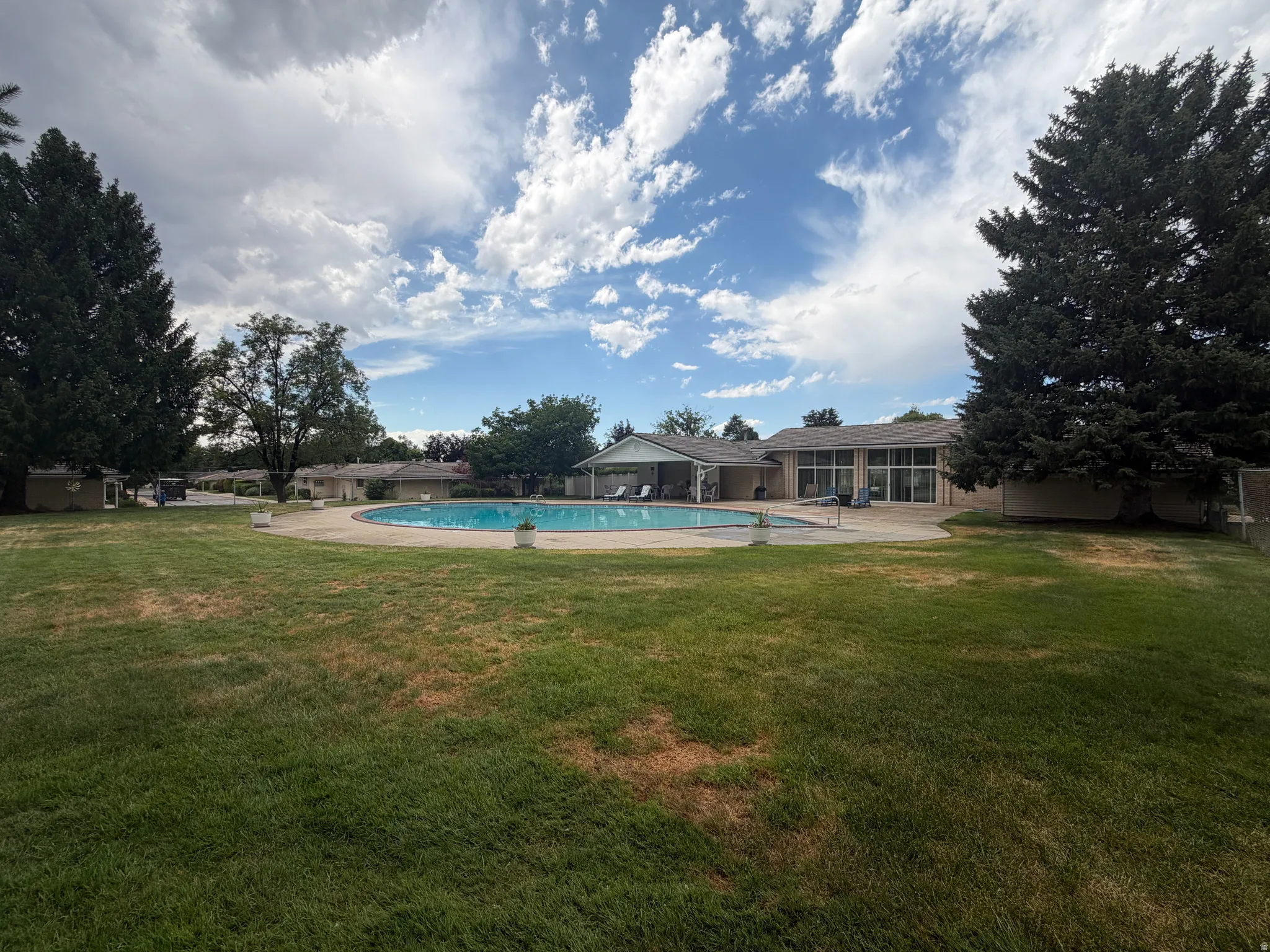 Club House Expansive lawn area fronting a large swimming pool with surrounding patio, mature trees, and a single-story building featuring multiple windows