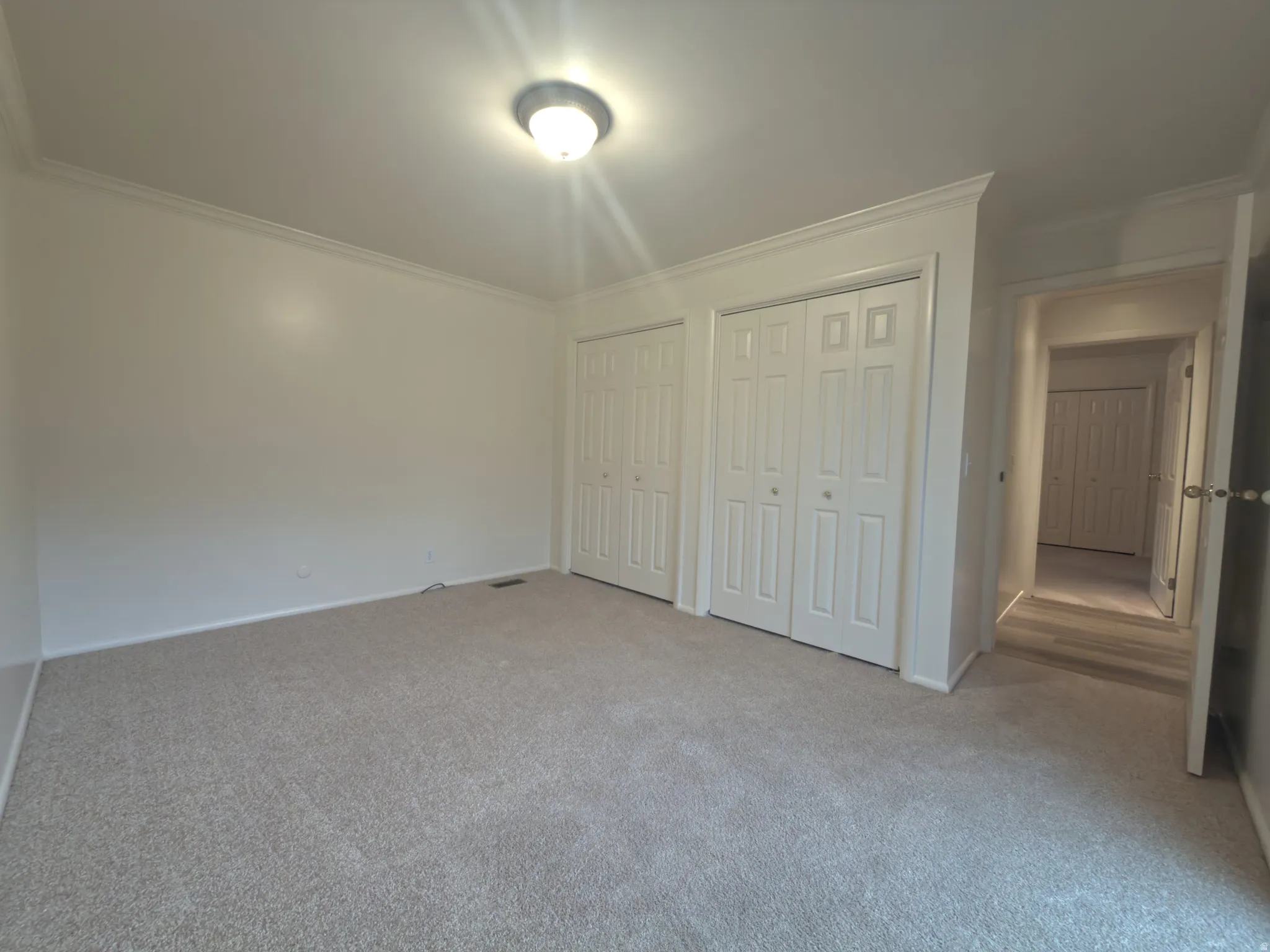Spacious room featuring neutral carpet flooring, a flush-mount ceiling light fixture, crown molding, and two sets of bi-fold closet doors