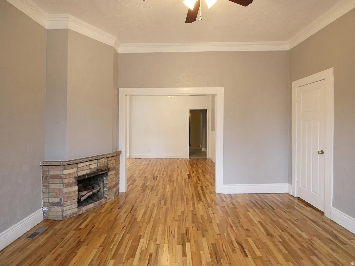 Spacious room featuring wood flooring, a brick fireplace, white baseboards, crown molding, and a ceiling fan