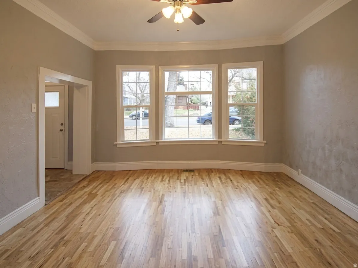 Hardwood flooring throughout this room features a ceiling fan with integrated lighting, a triple window assembly, baseboard trim, and crown molding